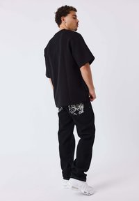 Zwart oversized T-shirt met korte mouwen, gecombineerd met zwarte jeans met witte bloemenpatronen en witte sneakers. Eenvoudig, modern ontwerp.