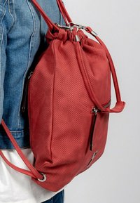 Sac à dos en cuir rouge avec texture perforée, fermeture à cordon, quincaillerie argentée et une poche extérieure zippée. Sangles latérales et bretelles visibles.