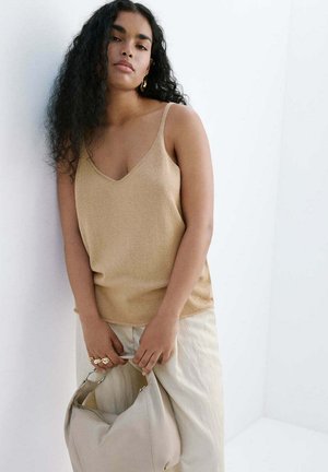Femme aux longs cheveux bouclés portant un haut beige sans manches et un pantalon clair, tenant un sac à main couleur crème sur fond blanc.
