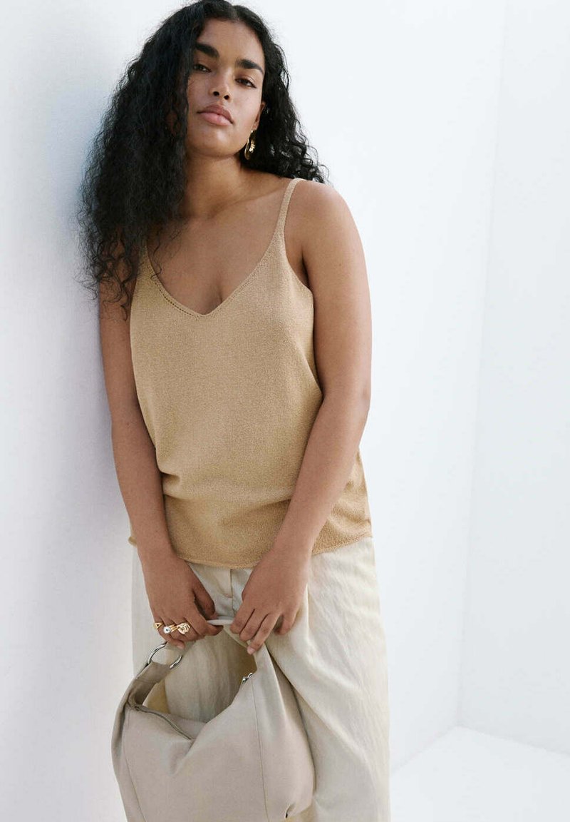 Femme aux longs cheveux bouclés portant un haut beige sans manches et un pantalon clair, tenant un sac à main couleur crème sur fond blanc.