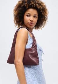 Νεαρή γυναίκα με σγουρά μαλλιά που φοράει ανοιχτό μπλε φόρεμα με λουλούδια και κρατάει σκούρη καφέ δερμάτινη τσάντα ώμου, κοιτάζοντας πλευρικά.