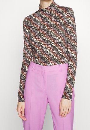 Vrouw met een lange mouw en een multicolor polkadotpatroon top, ingestopt in hooggetailleerde lavendelkleurige broek, tegen een witte achtergrond.