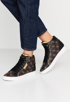 Piernas con vaqueros oscuros que llevan zapatillas altas negras y marrones con detalles dorados y suelas blancas, situadas sobre un suelo blanco.