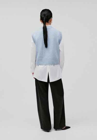 Mouwloos, lichtblauw gebreid vest met geribde zoom, gedragen over een wit blouse. Zwarte wijde broek en puntige zwarte schoenen maken de look af.