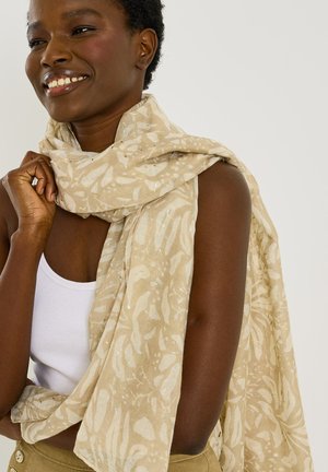 Mujer sonriente con el cabello corto y rizado se envuelve una bufanda beige con estampado alrededor de los hombros, vistiendo una camiseta sin mangas blanca y pantalones caqui.