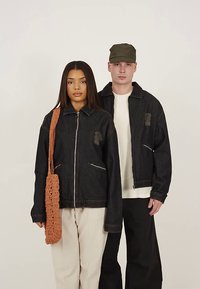 Dark denim jacket with two zippered pockets, worn by two models. One model carries a crocheted brown bag, and the other wears a cap.