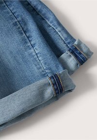 Revers de manches retroussés d'un jean en denim bleu clair montrant des coutures piquées et des bandes de tissu bleu foncé contrastantes sur un fond uni.
