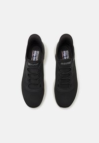 Baskets de sport noires avec tige en tissu en maille, lacets élastiques et semelle en caoutchouc blanche. Présente un logo sur la languette et un design flexible.