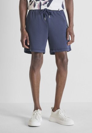 Hombre vistiendo pantalones cortos azul marino con cordón y zapatillas blancas, de pie contra un fondo claro y liso.