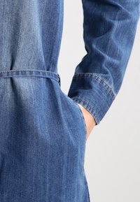 Sininen farkkutakki, jossa on mukautettu siluetti, pitkät hihat ja huomattavaa sauman yksityiskohtaa. Sisältää vyöllisen vyötärön ja sivutaskut.