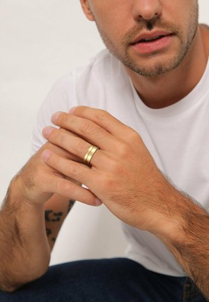 Gold-colored ring with two parallel bands, worn on a hand. The fingers have a natural, unpolished look, and the background is neutral.