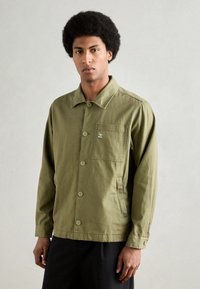 Olive green jacket made of cotton, featuring a button-up front, oval collar, and a chest pocket with a logo. Long sleeves and a relaxed fit.