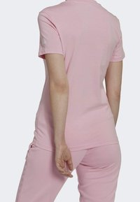 Světle růžová košile s krátkým rukávem, která má jednoduchý design. Tkanina vypadá měkce a hladce. Na rukávu je jemné logo.