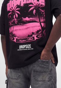 T-shirt noir avec un motif de plage tropicale rose, des palmiers, et le texte "Vacation Mode" et "Dropsize Designed in House// Since 2020."