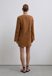 Brun ruskindjakke med lange ærmer og lige snit, der har minimal syning og en detalje med bagsøm. Båret over stroppede sandaler.