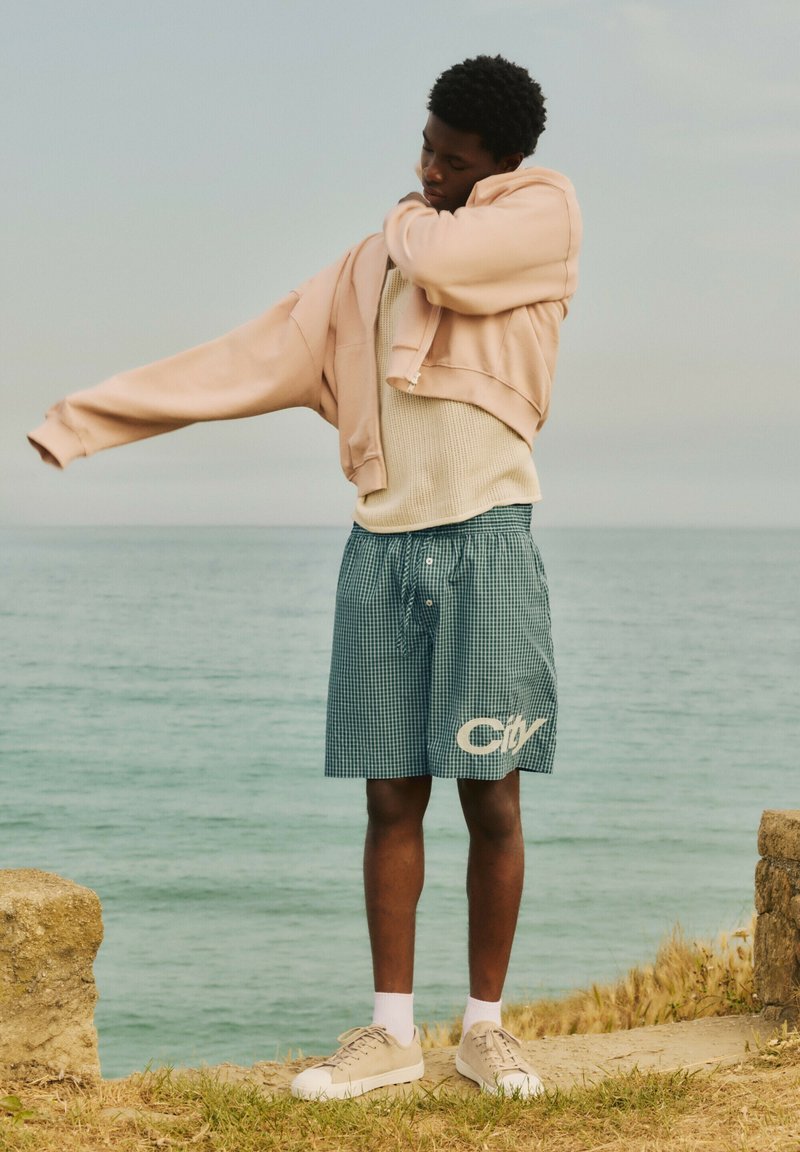 Jeune personne portant un short et des baskets enfile une veste beige, debout sur une falaise herbeuse au bord de la mer par une journée nuageuse.