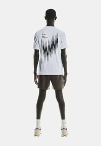 Mann steht mit dem R�ücken zur Kamera und trägt ein weißes T-Shirt mit schwarzem grafischen Muster und Text, braune Shorts, weiße Socken und beigefarbene Sneakers.