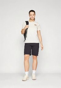 Light beige t-shirt with a small brown logo, paired with black shorts. Model wears white socks and beige sneakers, holding a black backpack.