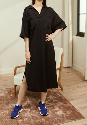 Kobieta stojąca w pomieszczeniu, ubrana w luźną czarną sukienkę midi z bocznymi rozcięciami i niebieskie trampki na brązowym dywanie obok drewnianego krzesła.