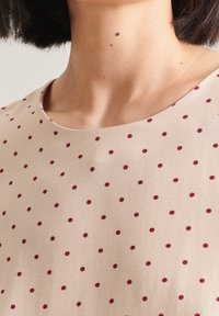 Blusa in tessuto beige con scollatura rotonda e un motivo di piccoli pois color mogano, con una texture liscia.