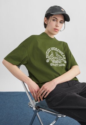 Person iført en grøn "Dickies Workwear" T-shirt og sort kasket, siddende på en metalstol med blå tæppe og grå væg i baggrunden.