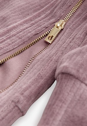 Svijetloružičasta jakna sa patentnim zatvaračem, izrađena od teksturiranog materijala, s zlatnim patentnim zatvaračem i glatkom podstavom. Detalj patentnog zatvarača i sastava tkanine vidljiv.