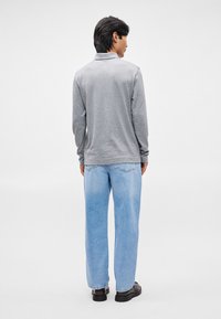 Polo grigio a maniche lunghe con vestibilità rilassata, abbinato a jeans azzurri larghi e scarpe nere, mostrato da dietro su uno sfondo semplice.