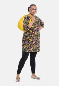 Camouflage tuniek in groen, paars en geel, gecombineerd met zwarte leggings en metallic platte schoenen; geaccessoireerd met een gele handtas.