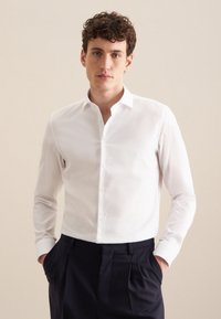 Camicia bianca a maniche lunghe in cotone con colletto stretto e chiusura frontale con bottoni, caratterizzata da un tessuto liscio e strutturato. Abbinata a pantaloni scuri.