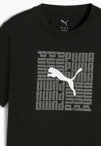 Melns kokvilnas T-krekls ar lielu baltu Puma logo un atkārtotu tekstu "PUMA" dažādās pelēkās nokrāsās.