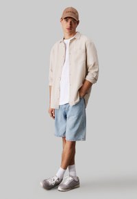 Camisa de botones color beige claro, camiseta blanca, pantalones cortos de mezclilla azul claro, zapatillas grises, calcetines blancos y una gorra color marrón claro con logotipo.