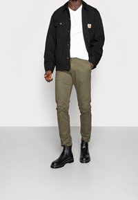 Man som bär svart jacka, vit t-shirt, olivgröna byxor och svarta läderstövlar, står mot en slät vägg.
