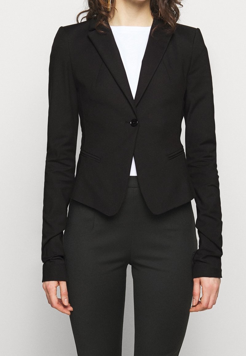 Femme portant un blazer noir cintré avec un seul bouton sur un haut blanc et un pantalon noir, debout devant un fond uni.