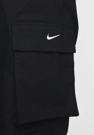 Melnas kravnesošās šortas, izgatavotas no teksturēta auduma, ar lielu sānu kabatu un baltu Nike logo priekšpusē.