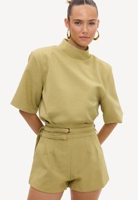 Femme portant un short taille haute ceinturé vert olive clair et un haut à manches courtes assorti avec col montant, debout sur fond blanc.