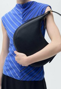 Black leather handbag with curved design, held under a blue and white striped sleeveless top. Smooth finish and minimal hardware details.