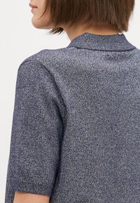 Kortermet strikket topp i mørkeblå med en subtil glans. Har en klassisk krage og teksturert stoff, som viser en tettsittende design.