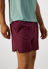 Mann in bordeauxroten Sportshorts mit seitlichen Taschen und einem mintgrünen Kurzarmshirt, steht vor einem neutralen Hintergrund.
