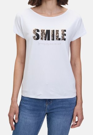 Kobieta nosząca białą koszulkę z krótkim rękawem z napisem "SMILE" w panterkę oraz ciemnoniebieskie dżinsy.