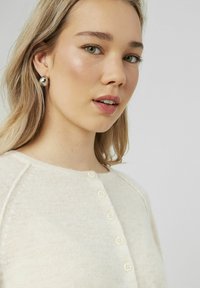 Cardigan en tricot crème avec un col rond, doté de cinq boutons décoratifs. Le mannequin porte des boucles d'oreilles métalliques surdimensionnées. Texture douce mise en valeur.