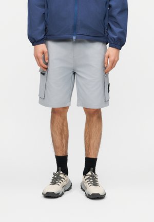 Mężczyzna w jasnoszarych szortach typu cargo, czarnych skarpetkach, beżowych butach trekkingowych i granatowej kurtce przeciwwiatrowej.