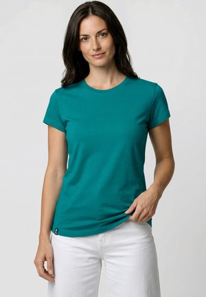 Frau mit dunklem Haar, die ein kurzärmliges türkisfarbenes T-Shirt und weiße Hosen trägt, vor einem schlichten hellgrauen Hintergrund stehend.