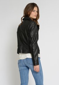Svart lädermotorcykeljacka med en figursydd design, slät textur och dragkedjestängning, bärs över en vit blus och blå jeans.