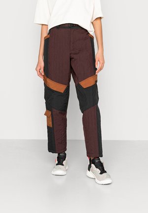 Personne portant un pantalon cargo bicolore marron, noir et rouille avec une chemise blanche et des baskets blanches et noires, debout sur un sol gris.