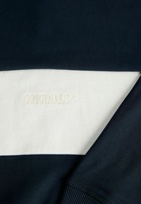 Gros plan d’un tissu rayé bleu marine et blanc avec le mot « ORIGINALS » brodé en fil blanc sur la rayure blanche.