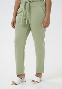 Pantalons taillés vert clair avec passants de ceinture, poches latérales et texture lisse. Associés à des sandales à talons blanches avec un design à bout ouvert.