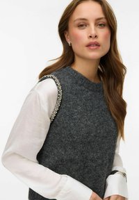 Sivi vuneni prsluk s okruglim izrezom i ukrasima uz otvor za ruku, slojevit preko bijele košulje s dugim rukavima, s glatkom teksturom.