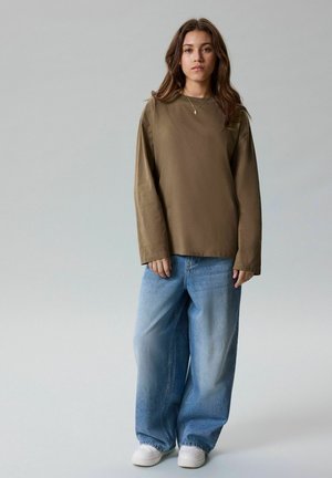 Jonge vrouw die een oversized bruin shirt met lange mouwen, losse blauwe spijkerbroek en witte sneakers draagt, staand tegen een effen lichtgrijze achtergrond.