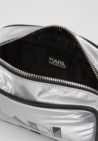 Silberne Nylon-Mäppchen mit schwarzen Akzenten, ausgestattet mit einem oberen Reißverschluss und einem Innenetikett mit der Aufschrift „KARL LAGERFELD“. Strukturierte Innenauskleidung sichtbar.