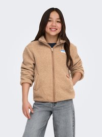 Teenagepige med langt mørkt hår iført en beige fleecetrøje med lynlås og lyseblå jeans, smilende med den ene hånd i lommen.